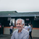 Portrait of a senior man with a beard smiling warmly outdoors wearing a plaid shirt.