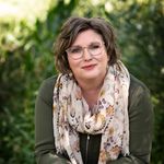 Portrait of a smiling woman wearing eyeglasses and a floral scarf, leaning on a wooden railing outdoors.